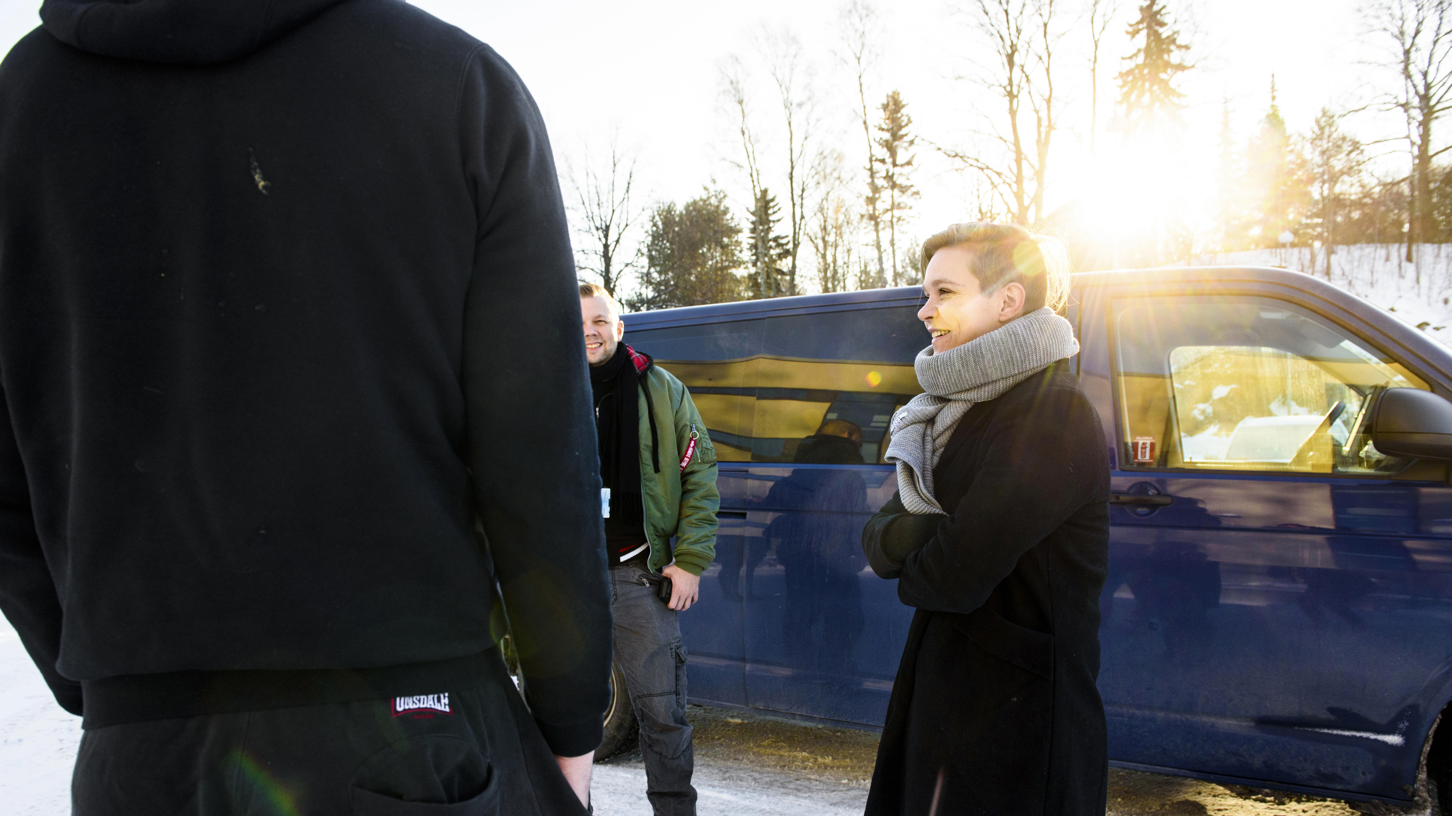 A woman and a man talk with another man, who is facing away from the camera, in front of a blue van outdoors in winter.