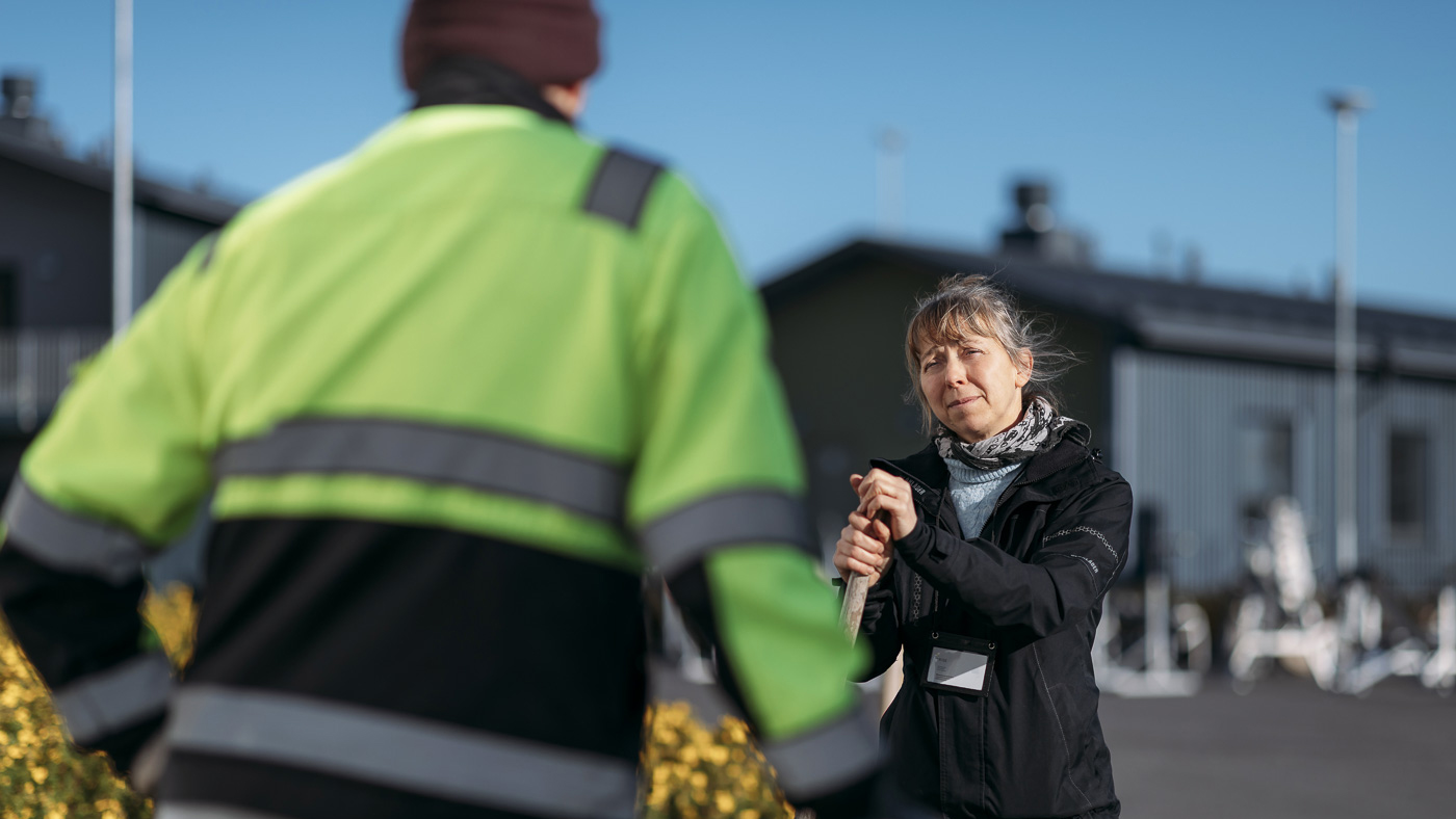 A man wearing a high-visibility jacket talks with a woman. The woman leans on a broom handle.