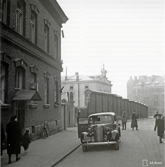 Van­ha mus­ta­val­koi­nen kuva kau­pun­gin ka­dul­ta, jol­la ovi auki ole­va auto sei­soo kor­kean ra­ken­nuk­sen vie­res­sä. Kadulla kävelee ihmisiä.