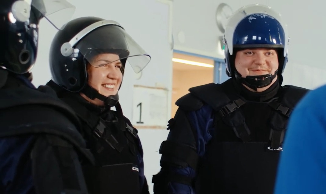 Two people wearing bullet-proof vests and protective helmets are talking with a third person who is facing away from the camera. One of the people visible in the picture has a grin on her face.